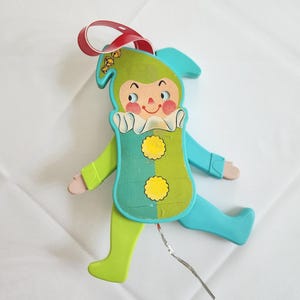 May include: A vintage, colorful wooden toy in the shape of a jester. The jester has a green hat and body, blue arms and legs, and a smiling face. It has a red ribbon for hanging and a silver wire extending from the body.