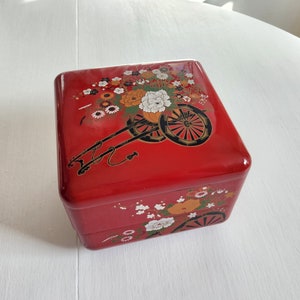 Puede incluir: Una caja de madera roja de dos niveles con un diseño floral. El nivel superior presenta un carro negro y dorado con flores. El nivel inferior tiene un diseño floral más pequeño.