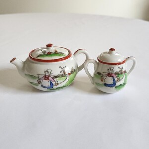 May include: A miniature ceramic teapot and sugar bowl set featuring hand-painted illustrations of a person in traditional clothing carrying buckets, a windmill, and green landscape scenery, accented with a thin red rim on the lids and bodies.