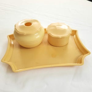 May include: A vintage vanity set in a soft yellow hue. The set includes a tray with a scalloped edge, a lidded jar, and a small container with a matching lid. The items are made of a smooth, possibly plastic material.