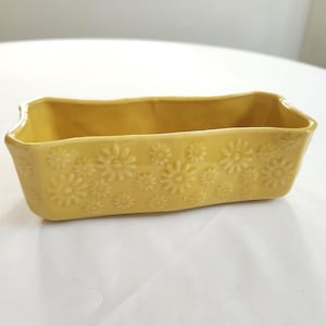 May include: A rectangular, yellow ceramic dish with a floral pattern. The dish has a slightly wavy rim and is likely used for baking or serving. The daisy-like flowers are embossed on the sides of the dish.