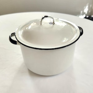 May include: White enamel pot with a lid and black trim. The pot has two black handles and a silver handle on the lid. The pot is sitting on a white surface.