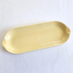May include: A pale yellow ceramic serving tray with an elongated oval shape, featuring a smooth, glossy finish and subtle concentric ring detailing along the raised edges, designed with small integrated handles on both ends of the dish.