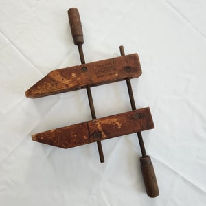 May include: A vintage Jorgensen wood clamp with a dark brown finish. The clamp features a wooden frame, metal screws, and wooden handles. The text "TRUE MARK JORGENSEN REG. U.S. PAT. OFF. MADE IN U.S.A." is visible on the wood.