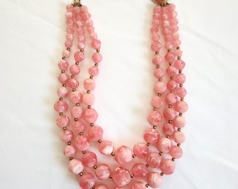May include: A three-strand pink beaded necklace with a matching round clasp. The beads are a marbled pink and white, with small gold-colored spacers. The necklace is displayed on a white background.