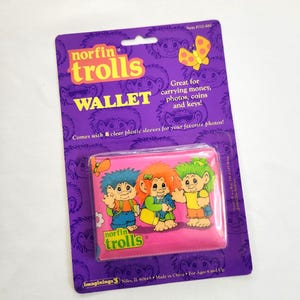 May include: A vintage "Norfin Trolls" wallet on a purple backing. The pink wallet features three cartoon trolls and the text "Norfin Trolls." The packaging states it's great for carrying money, photos, coins, and keys.