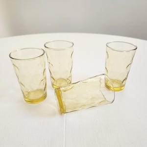May include: Four vintage yellow-tinted drinking glasses. Three glasses are upright, and one is lying on its side. The glasses have a textured, dimpled surface and a rounded base. The glasses are made of glass.