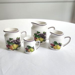 May include: Four white ceramic pitchers with a fruit design. Each pitcher has a handle and a dark purple rim. The fruit design includes grapes, pears, and other colorful fruits.