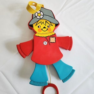 May include: A vintage pull-string toy scarecrow with a red shirt, blue pants, and a gray hat. The toy has a yellow pull string and a red ring.
