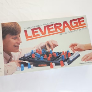 May include: A game box for the board game Leverage. The box is yellow with red and black text. The game is for 2 players and is a game of strategy and suspense. The box shows a game board with red and blue pieces. The text on the box says "Don't get caught off balance!"
