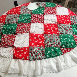 May include: A round Christmas tree skirt with a patchwork design in red, green, and white fabrics. The skirt has a ruffled white eyelet lace trim. The patchwork features floral and dotted patterns.