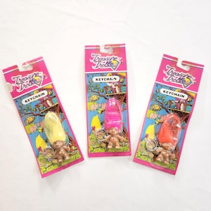 Puede incluir: Tres llaveros de Treasure Trolls en embalaje rosa. Cada llavero presenta una peque&ntilde;a mu&ntilde;eca troll con diferentes colores de pelo: amarillo, rosa y naranja. El embalaje incluye el texto "Treasure Trolls" y "KEYCHAIN".