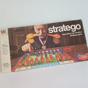 May include: A vintage board game box for the game Stratego. The box features a man in a military uniform with a mustache and a medal. The game is described as a "fascinating two-handed strategy game".