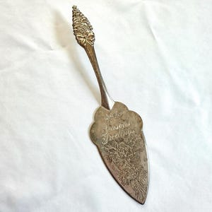 May include: Silver cake server with a decorative handle and a floral design on the blade. The blade is engraved with the words "Seasons Greetings". The handle has a detailed, textured design.
