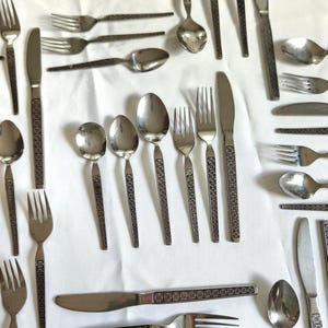May include: An assortment of vintage stainless steel flatware, including forks, knives, and spoons. The handles feature a decorative, geometric pattern. The silverware is arranged on a white surface, showcasing the variety of pieces.