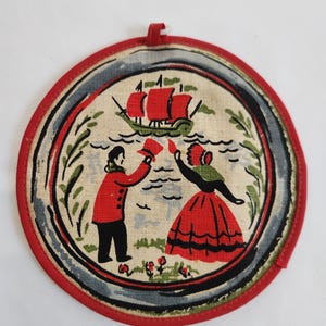 May include: Round red and white fabric pot holder with a blue and green border. The design features a couple in red and black clothing standing by a body of water with a red sailboat in the background.