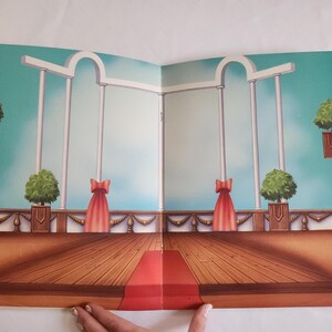 Puede incluir: Un tel&oacute;n de fondo de papel impreso que muestra una escena de sal&oacute;n de baile con suelo de madera, una alfombra roja central, columnas arquitect&oacute;nicas blancas y plantas verdes. La escena incluye cortinas rojas y un cielo azul claro.