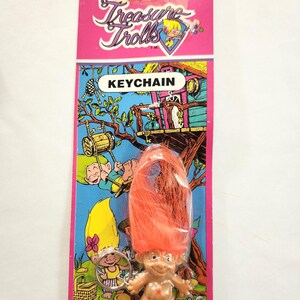 Puede incluir: Un llavero vintage de Treasure Trolls en una tarjeta rosa y azul. El llavero presenta una peque&ntilde;a figura de troll con pelo naranja. La tarjeta tiene las palabras "Treasure Trolls" y "KEYCHAIN" impresas, junto con ilustraciones de dibujos animados.