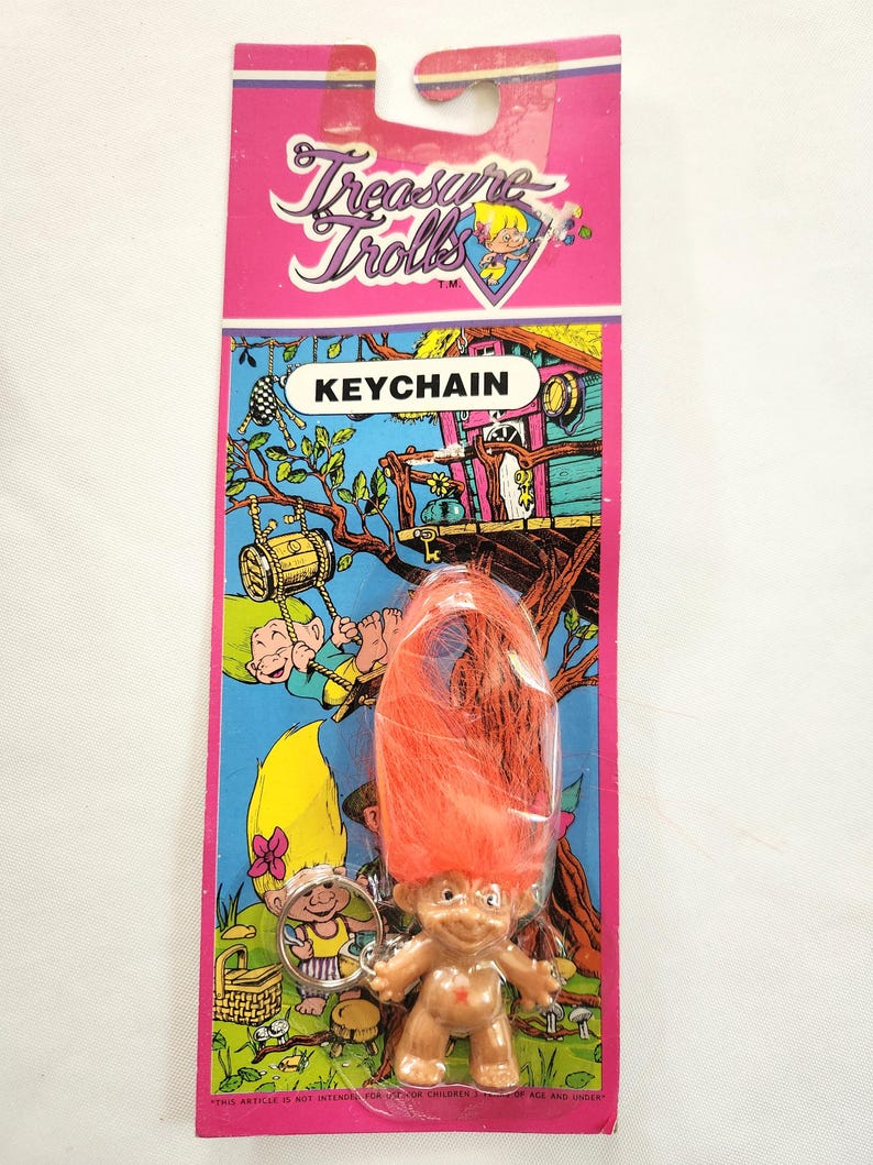 Puede incluir: Un llavero vintage de Treasure Trolls en una tarjeta rosa y azul. El llavero presenta una peque&ntilde;a figura de troll con pelo naranja. La tarjeta tiene las palabras "Treasure Trolls" y "KEYCHAIN" impresas, junto con ilustraciones de dibujos animados.
