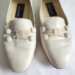 Puede incluir: Un par de mocasines de cuero blanquecino. Cada zapato presenta un lazo decorativo hecho de cord&oacute;n beige y tres adornos redondos blancos. El nombre de la marca est&aacute; impreso en una etiqueta negra dentro de cada zapato.