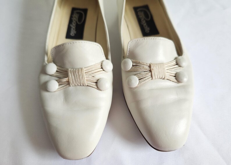 Puede incluir: Un par de mocasines de cuero blanquecino. Cada zapato presenta un lazo decorativo hecho de cord&oacute;n beige y tres adornos redondos blancos. El nombre de la marca est&aacute; impreso en una etiqueta negra dentro de cada zapato.