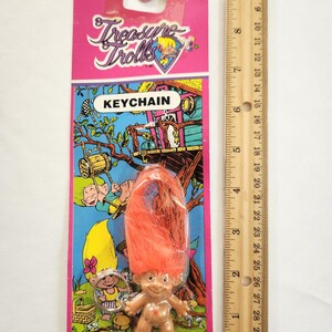 Puede incluir: Un llavero vintage de Treasure Trolls en una tarjeta rosa y azul. El llavero presenta una peque&ntilde;a figura de troll con pelo naranja. La tarjeta tiene el texto "Treasure Trolls" y "KEYCHAIN". Una regla mide hasta 25 cm.