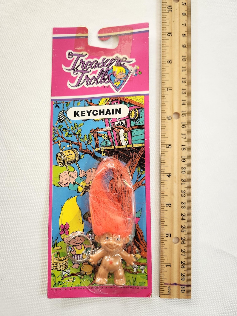 Puede incluir: Un llavero vintage de Treasure Trolls en una tarjeta rosa y azul. El llavero presenta una peque&ntilde;a figura de troll con pelo naranja. La tarjeta tiene el texto "Treasure Trolls" y "KEYCHAIN". Una regla mide hasta 25 cm.