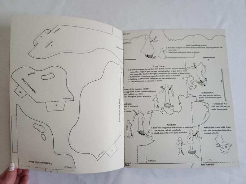 Puede incluir: Un libro de manualidades en papel con plantillas de arte lineal en blanco y negro de personajes de Disney, incluyendo al Rey Trit&oacute;n, el Pr&iacute;ncipe Eric, Sebasti&aacute;n y Ariel. Las p&aacute;ginas contienen instrucciones, l&iacute;neas de pliegue y formas.