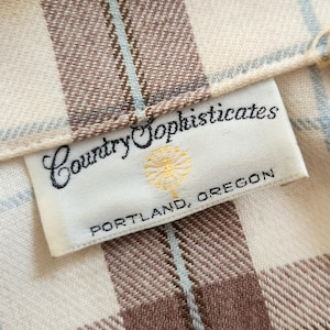 May include: Close-up of a fabric with a plaid pattern in cream, brown, and light blue. The label reads "Country Sophisticates, Portland, Oregon."
