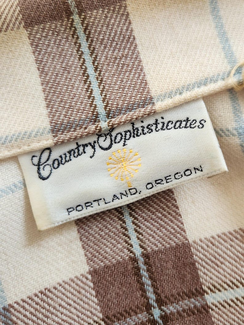 May include: Close-up of a fabric with a plaid pattern in cream, brown, and light blue. The label reads "Country Sophisticates, Portland, Oregon."