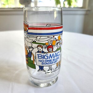May include: A clear glass with a blue, red, and white stripe design. The glass features a cartoon illustration of people holding signs that say "Big Mac" and "No one else stacks up".