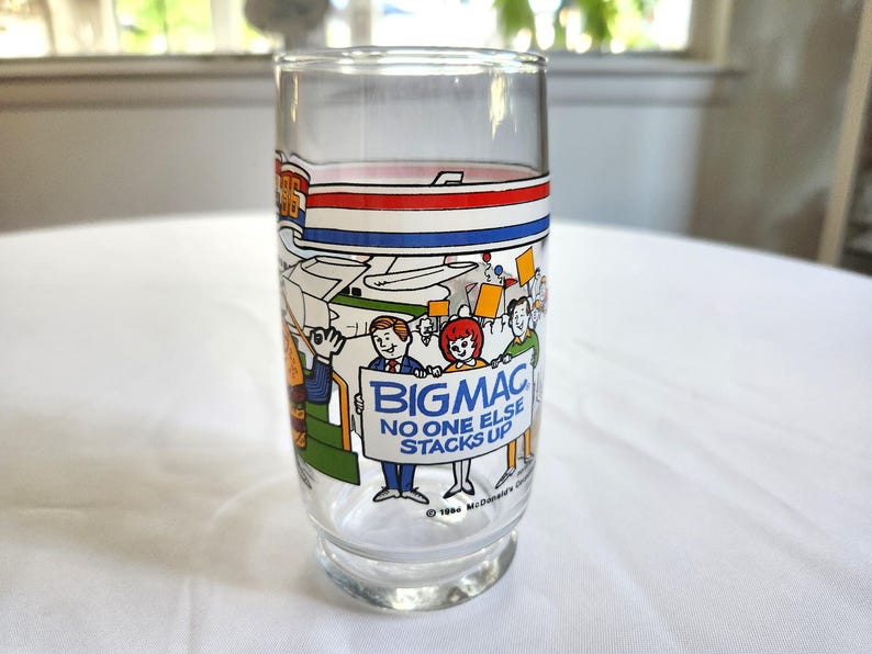 May include: A clear glass with a blue, red, and white stripe design. The glass features a cartoon illustration of people holding signs that say "Big Mac" and "No one else stacks up".