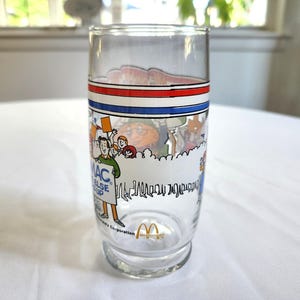 May include: A clear glass with a red, white, and blue band around the top. The glass has a cartoon image of people holding a sign that says "Mac, I'd rather have you than anything else in the world." The glass also has the McDonald's logo on the bottom.
