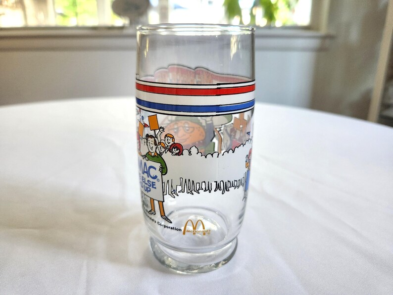 May include: A clear glass with a red, white, and blue band around the top. The glass has a cartoon image of people holding a sign that says "Mac, I'd rather have you than anything else in the world." The glass also has the McDonald's logo on the bottom.