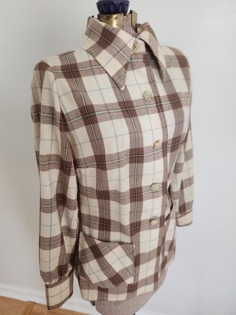 May include: A vintage plaid jacket in cream, brown, and light blue. The jacket features a collared neckline, button-down front, long sleeves, and two front pockets. The plaid pattern is a classic design.