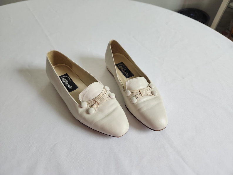 Puede incluir: Un par de mocasines de cuero color crema. Cada zapato presenta un lazo decorativo con tres peque&ntilde;os adornos redondos y una etiqueta de marca en el interior. Los zapatos est&aacute;n sobre un fondo blanco liso.