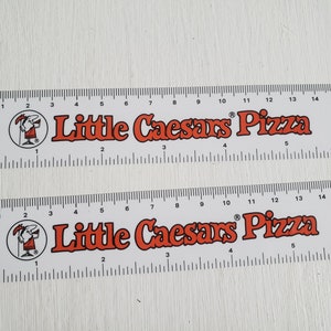 Puede incluir: Dos reglas de pl&aacute;stico blanco con marcas negras y texto rojo. Las reglas miden 15 pulgadas de largo y presentan el logotipo de Little Caesars Pizza y una mascota de dibujos animados. Las reglas est&aacute;n una al lado de la otra.
