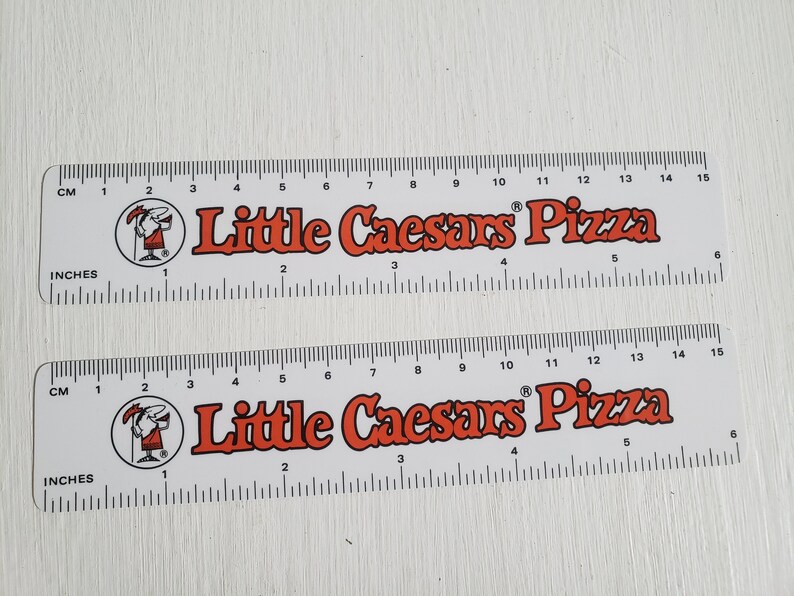 Puede incluir: Dos reglas de pl&aacute;stico blanco con marcas negras y texto rojo. Las reglas miden 15 pulgadas de largo y presentan el logotipo de Little Caesars Pizza y una mascota de dibujos animados. Las reglas est&aacute;n una al lado de la otra.