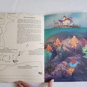 Puede incluir: Una p&aacute;gina doble de un libro de actividades de La Sirenita. La p&aacute;gina izquierda presenta plantillas de arte lineal en blanco y negro para manualidades con instrucciones. La derecha muestra una escena submarina colorida con peces.