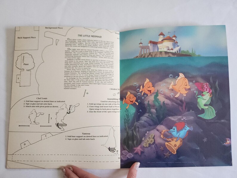 Puede incluir: Una p&aacute;gina doble de un libro de actividades de La Sirenita. La p&aacute;gina izquierda presenta plantillas de arte lineal en blanco y negro para manualidades con instrucciones. La derecha muestra una escena submarina colorida con peces.