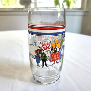 May include: A vintage glass with a red, white, and blue stripe design. The glass features a cartoon illustration of people holding signs that say "Vote for Mac" and "McDonald's".