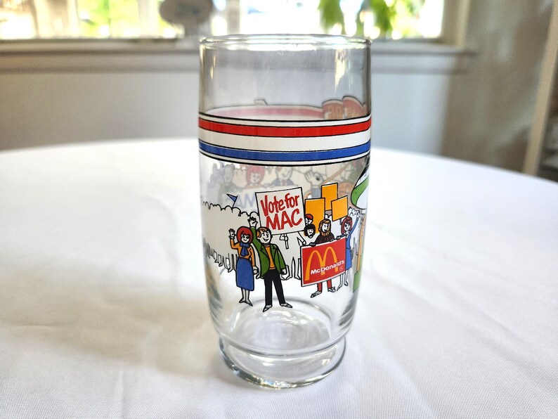 May include: A vintage glass with a red, white, and blue stripe design. The glass features a cartoon illustration of people holding signs that say "Vote for Mac" and "McDonald's".