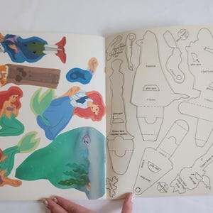 Puede incluir: Un libro de manualidades en papel con ilustraciones coloridas de personajes de La Sirenita en la p&aacute;gina izquierda y plantillas de recorte en blanco y negro con etiquetas como punto de pegamento en la p&aacute;gina derecha para el montaje.