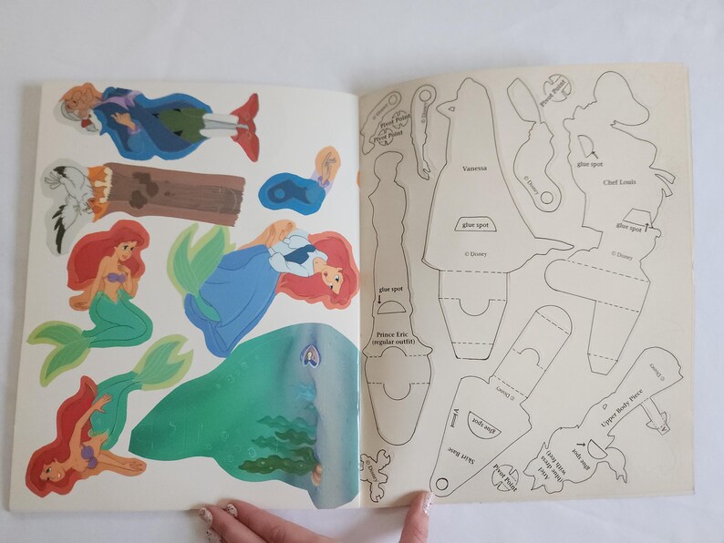 Puede incluir: Un libro de manualidades en papel con ilustraciones coloridas de personajes de La Sirenita en la p&aacute;gina izquierda y plantillas de recorte en blanco y negro con etiquetas como punto de pegamento en la p&aacute;gina derecha para el montaje.