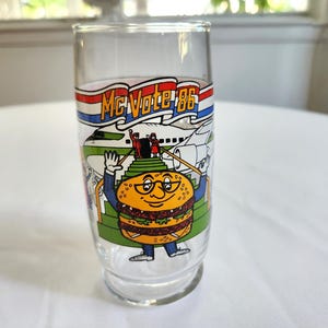 May include: A clear glass with a red, white, and blue banner that says "McVote '86". The glass features a cartoon character that looks like a hamburger with a face and glasses. The character is standing on a staircase with a plane in the background.