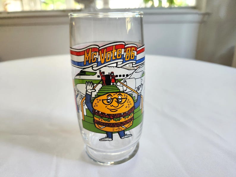 May include: A clear glass with a red, white, and blue banner that says "McVote '86". The glass features a cartoon character that looks like a hamburger with a face and glasses. The character is standing on a staircase with a plane in the background.
