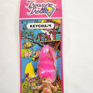 Puede incluir: Un llavero de Treasure Trolls en una tarjeta rosa y azul. El llavero presenta una peque&ntilde;a mu&ntilde;eca troll con pelo rosa. La tarjeta tiene las palabras "Treasure Trolls" y "KEYCHAIN" impresas, junto con ilustraciones de trolls.