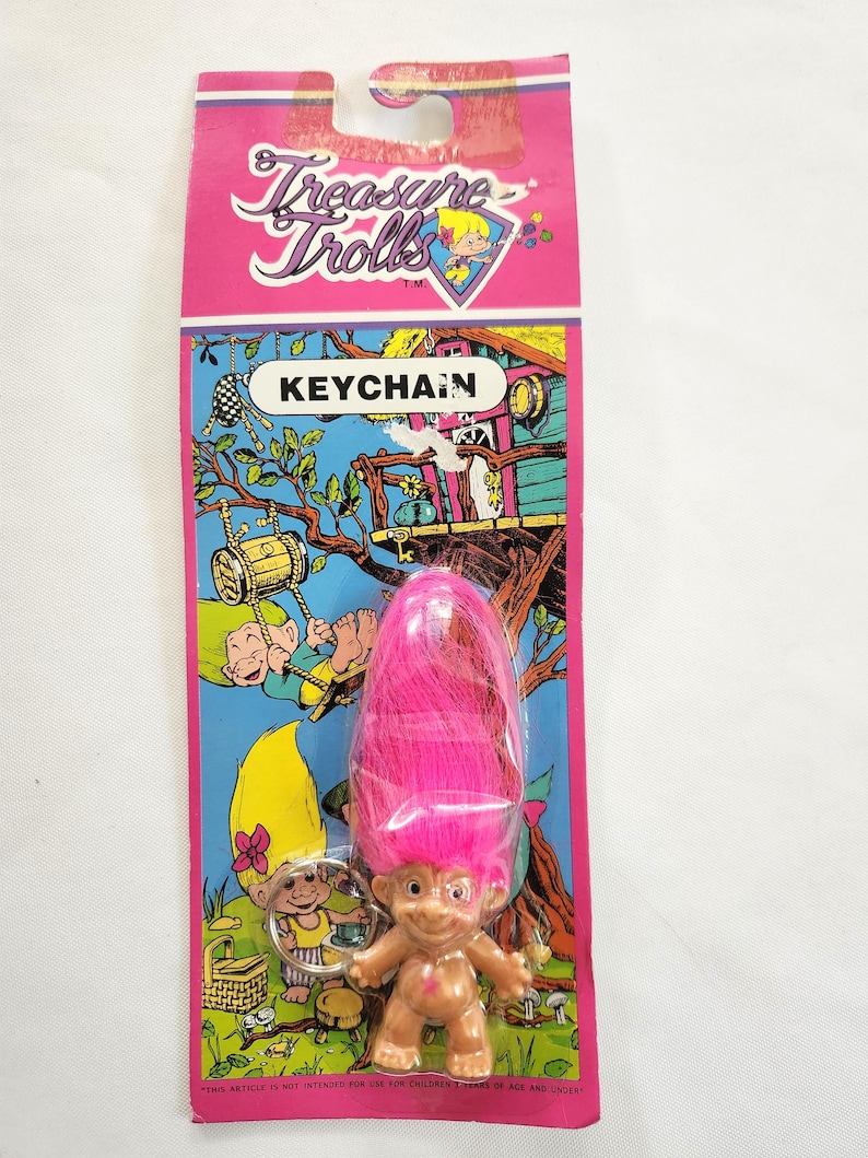 Puede incluir: Un llavero de Treasure Trolls en una tarjeta rosa y azul. El llavero presenta una peque&ntilde;a mu&ntilde;eca troll con pelo rosa. La tarjeta tiene las palabras "Treasure Trolls" y "KEYCHAIN" impresas, junto con ilustraciones de trolls.