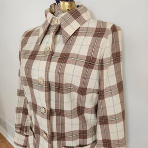 May include: A vintage plaid coat in cream, brown, and light blue. The coat features a collared neckline, button-down front, and two front pockets. The fabric has a classic plaid pattern, and the coat is displayed on a mannequin.