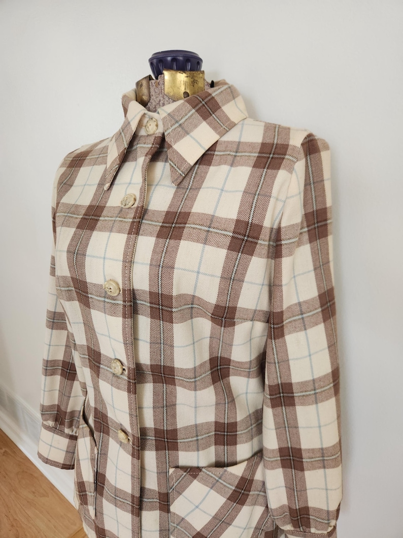 May include: A vintage plaid coat in cream, brown, and light blue. The coat features a collared neckline, button-down front, and two front pockets. The fabric has a classic plaid pattern, and the coat is displayed on a mannequin.